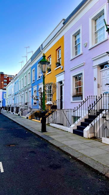 A row of colorful, multi-story terraced houses on a street in Notting Hill, London, with facades painted in shades of beige, dark blue, bright green, and pink. The buildings feature large windows, som