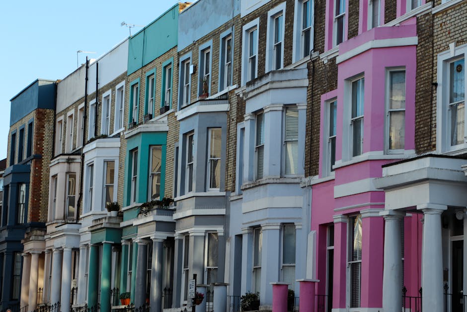 A row of Victorian terraced houses in Notting Hill featuring colorful facades in shades of blue, teal, white, and pink. Each property has a bay window with white trims, some decorated with potted plan
