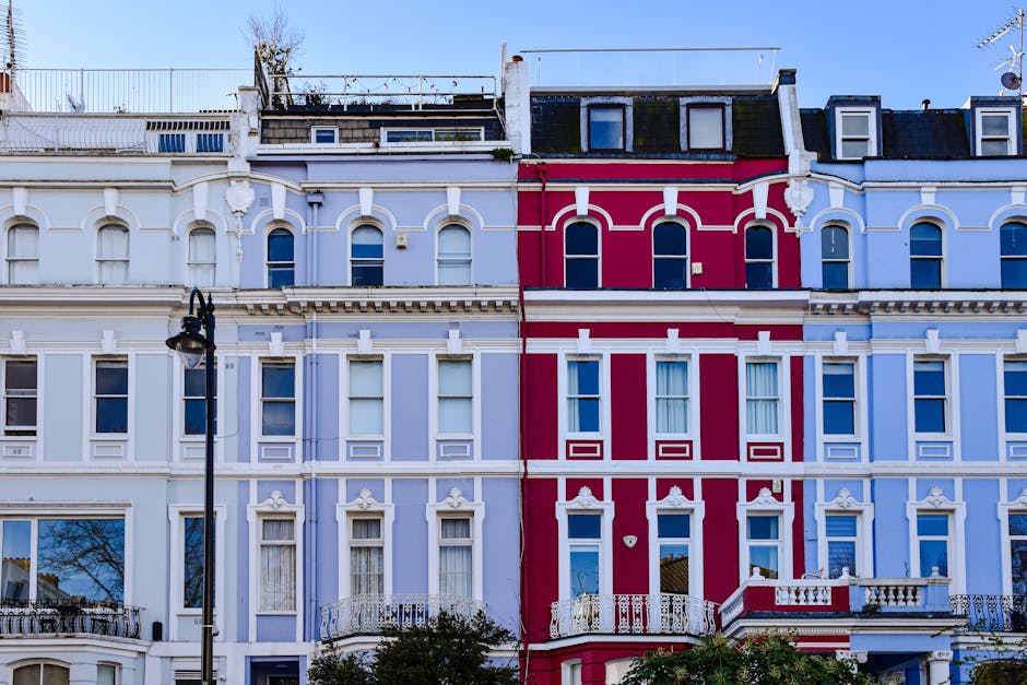 A row of Victorian terraced houses in Notting Hill featuring colorful facades in shades of blue, teal, white, and pink. Each property has a bay window with white trims, some decorated with potted plan